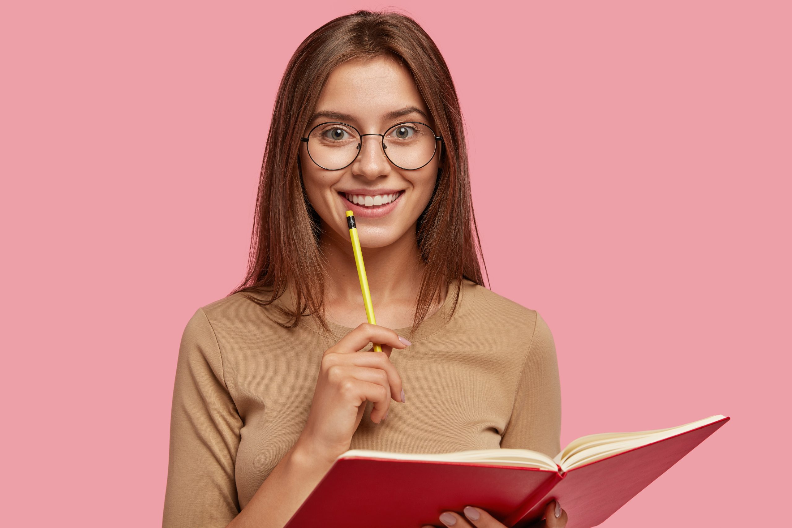 Photo of happy brunette woman with positive smile, carries textbook, holds pencil for writing, makes notes while listens some information, poses over pink background